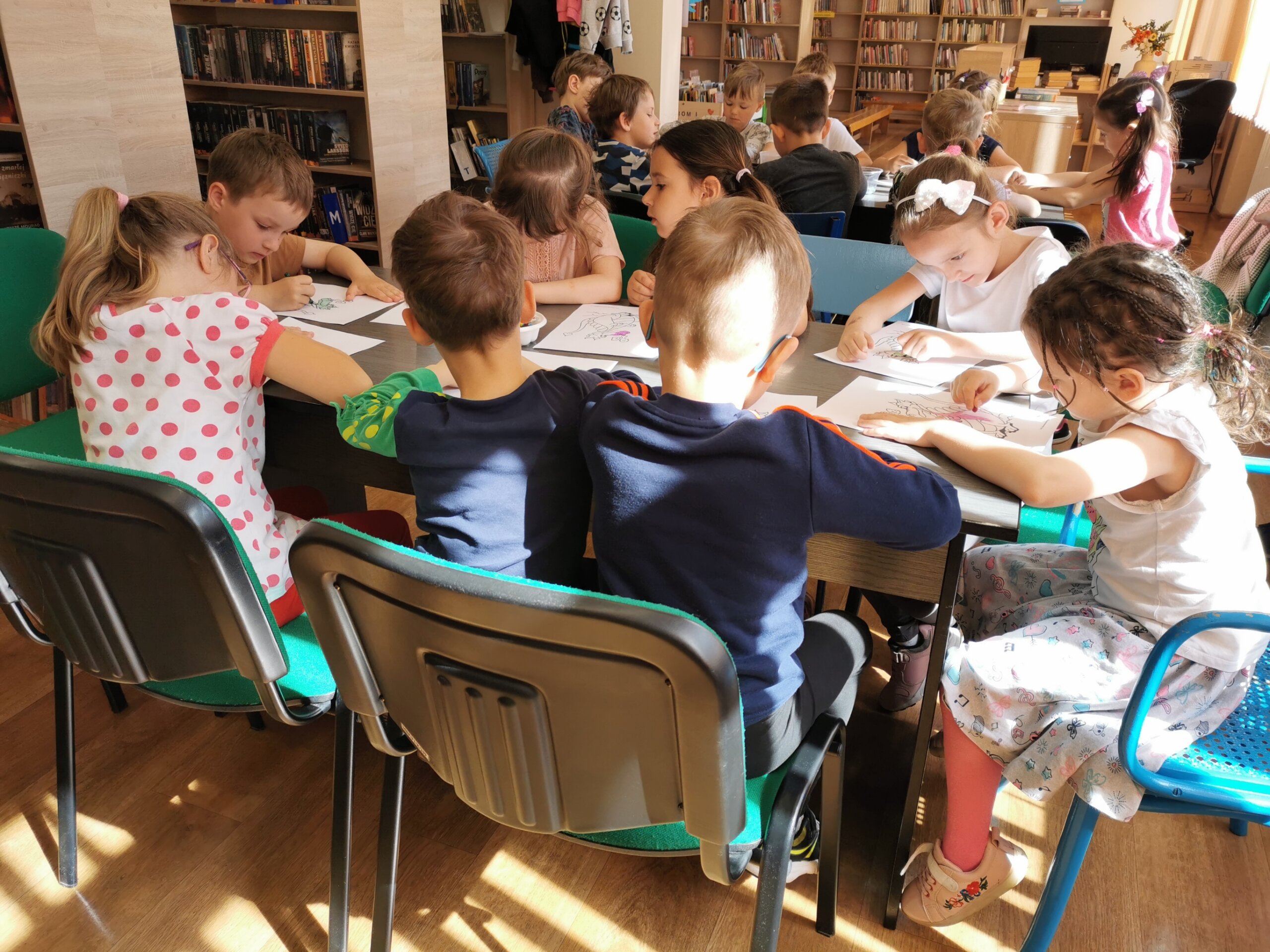 Dzieci w bibliotece siedzą przy stolikach i rysują