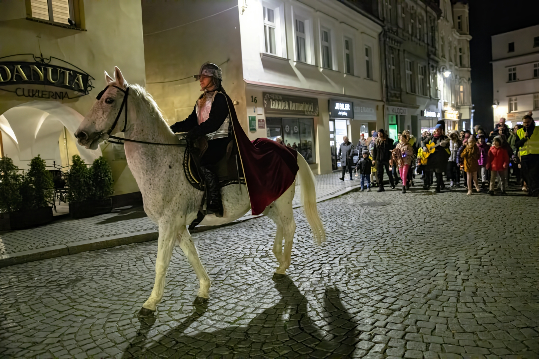 Aktorka przebrana za św. Marcina jedzie na koniu ulicą. Za nią idzie tłum ludzi.